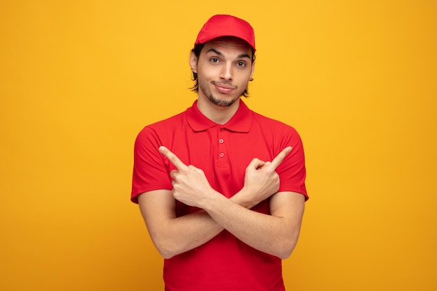 Zufriedener junger lieferbote in uniform und mütze, der in die kamera blickt und mit dem finger auf die seiten zeigt, die auf gelbem hintergrund isoliert sind