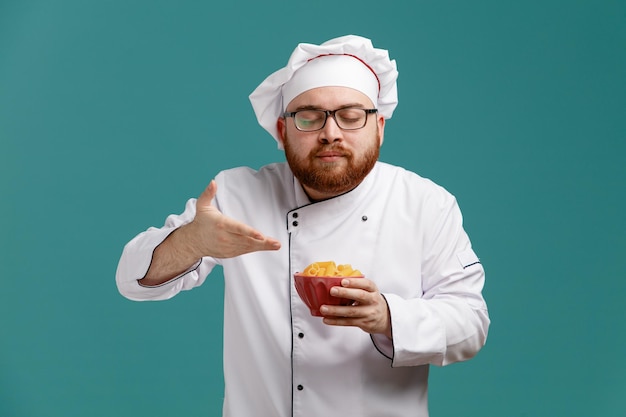 Zufriedener junger männlicher Koch mit Brille, Uniform und Mütze, die eine Schüssel mit Makkaroni hält, die mit der Hand darauf zeigt, Aromen von Essen riechend, mit geschlossenen Augen, isoliert auf blauem Hintergrund