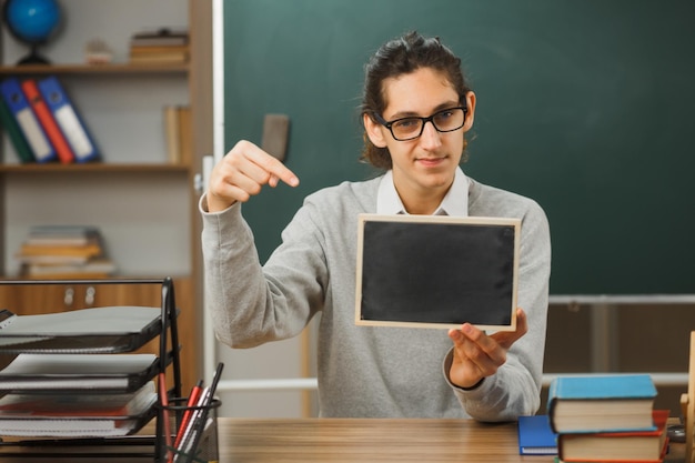 zufriedener junger männlicher lehrer mit brille, der am schreibtisch sitzt und auf eine mini-tafel mit schulwerkzeugen im klassenzimmer zeigt