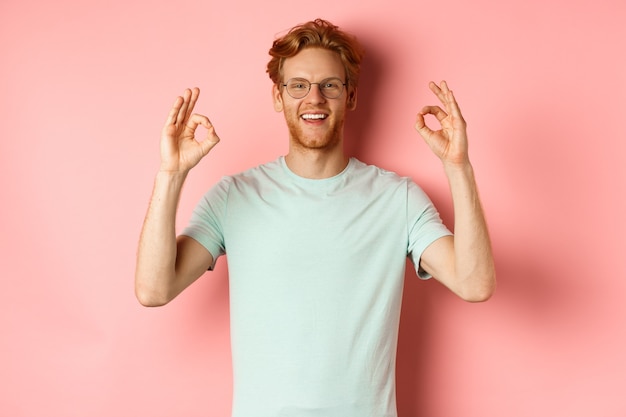 Zufriedener kaukasischer Mann mit roten Haaren mit Brille und T-Shirt, der in Ordnung Gesten zeigt und lächelt.