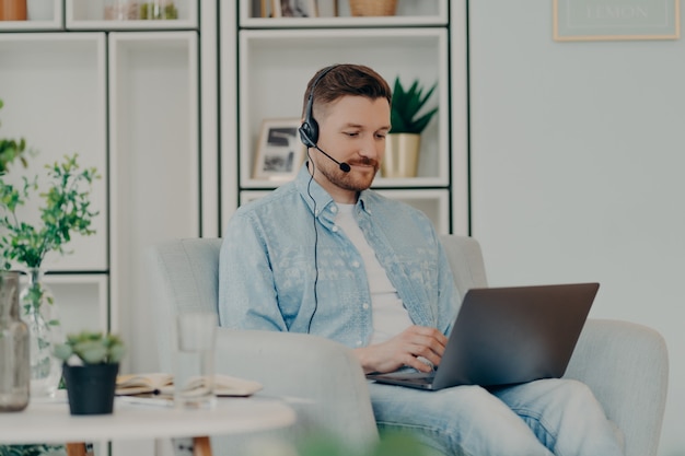 Zufriedener Kerl in bequemer Kleidung, der ein Headset für die Videokommunikation verwendet und den Laptop auf seinem Schoß hält, während er zu Hause bleibt. Männlicher Freiberufler, der online arbeitet. Fernarbeit und freiberufliches Konzept