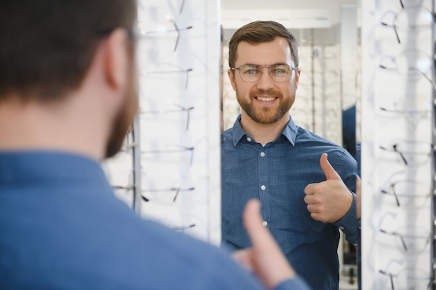 Zufriedener Kunde Ansicht eines glücklichen jungen männlichen Kunden, der eine neue Brille trägt, die in der Nähe von Rack und Vitrine mit Brillen steht Lächelnder Mann, der eine Brille anprobiert