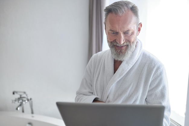 Zufriedener Mann mit dem Laptop, der im Badezimmer steht