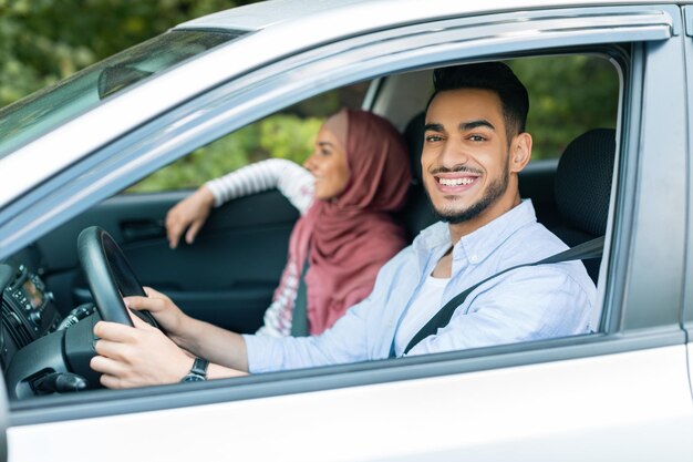 Zufriedener tausendjähriger arabischer Mann beim Fahren schaut sich Kamerafahrten mit seiner Frau im Hijab im Auto an und genießt die Fahrt