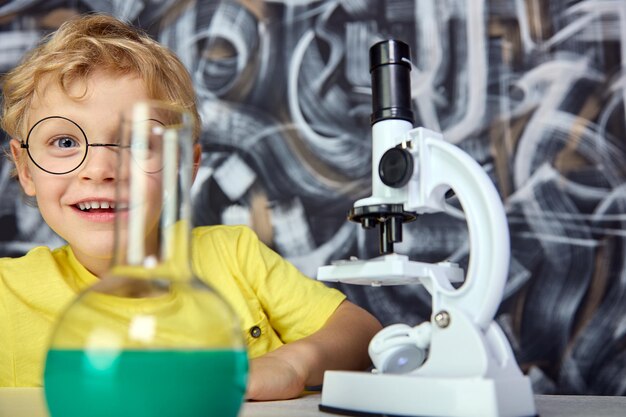 Zufriedenes Kind in runder Brille und T-Shirt versteckt sich hinter Laborgegenständen