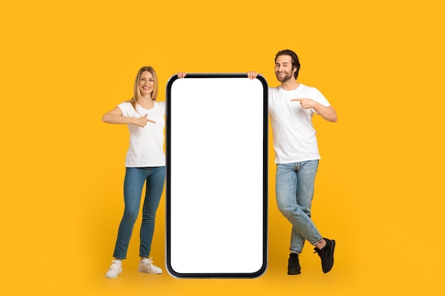 Zufriedenes tausendjähriges europäisches Paar in weißen T-Shirts zeigt mit dem Finger auf ein riesiges Telefon mit leerem Bildschirm