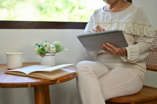 Zugeschnittenes Bild Asiatin im Alter der 60er Jahre, die am Fenster sitzt und ein tragbares digitales Tablet verwendet