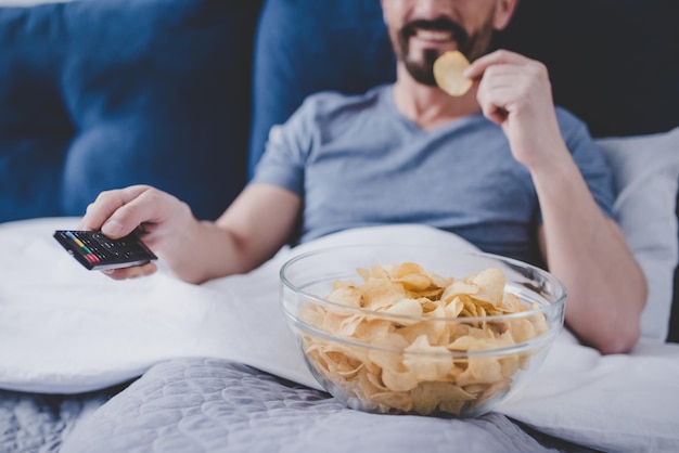 Zugeschnittenes Bild eines lächelnden Mannes, der fernsieht und Chips isst, während er sich zu Hause im Bett ausruht