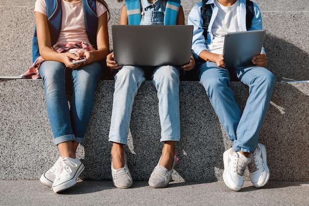 Zugeschnittenes Bild von Schulkindern, die draußen auf der Straße sitzen und digitale Geräte benutzen