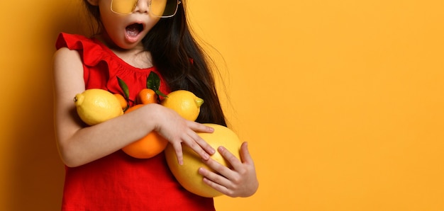 Zugeschnittenes Foto eines aufgeregten kleinen asiatischen Mädchens in einer roten Bluse, das Pomelo, Orangen und Zitronen in den Händen hält und auf einem orangefarbenen Hintergrund posiert. Kindheit, Früchte. Kopierbereich schließen