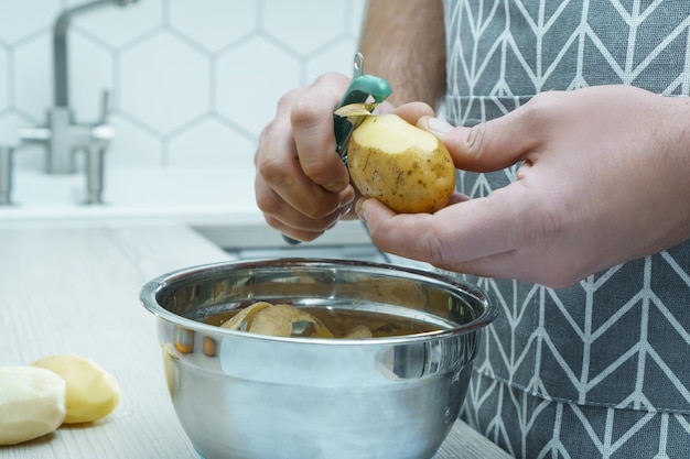 Zugeschnittenes Foto eines Mannes mit grauer Schürze, der Kartoffeln mit grünem Schäler über einer Metallschüssel schält Organische Abfallwirtschaft