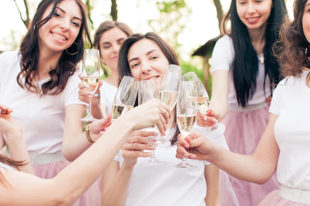 Zugeschnittenes Lichtfoto von glücklichen Mädchen, die auf der Junggesellenparty im Freien feiern, die Braut steht in der Mitte. Mädchen, die Gläser Champagner halten, klirren sie zusammen, lächeln und jubeln