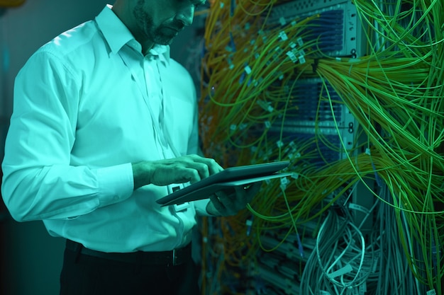 Zugeschnittenes Porträt eines gutaussehenden Dateningenieurs mit digitalem Tablet im Serverraum, während er mit Supercomputer in blauem Licht arbeitet, Kopierraum