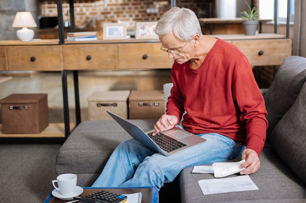 Zuhause arbeiten. Netter angenehmer älterer Mann, der einen Laptop hält und einen Knopf darauf drückt, während er zu Hause arbeitet