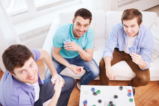 Zuhause, Freizeit, Spiele, Freundschaft, Glücksspiel und Unterhaltung - drei lächelnde männliche Freunde, die zu Hause Karten spielen