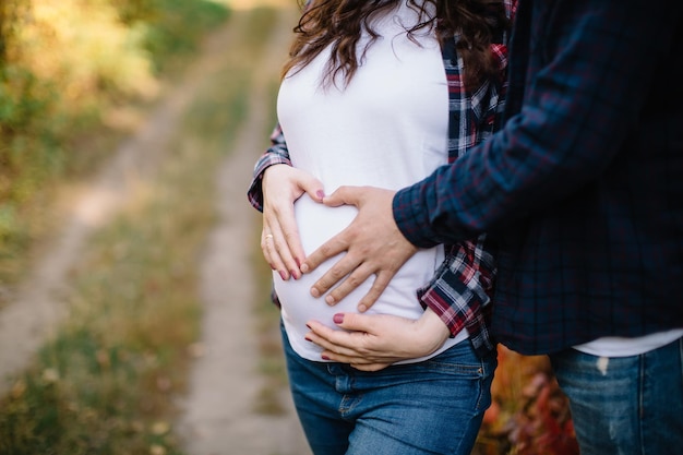 Zukünftige Eltern außerhalb der Stadt stehen und umarmen sich. Hände in Herzform auf dem Bauch der schwangeren Frau.