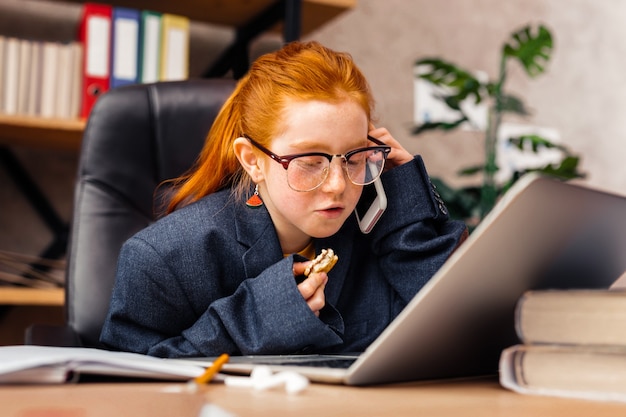 Zukünftige Geschäftsfrau. Nettes ernstes Mädchen, das am Telefon spricht, während es vor dem Laptop sitzt