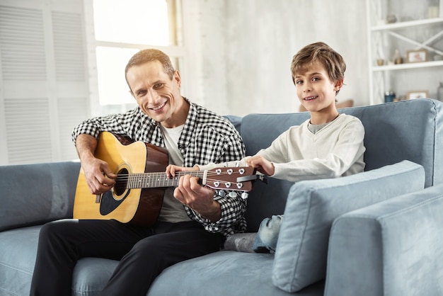 Zukünftiger Gitarrist. Gut aussehender, gut gebauter Mann, der die Gitarre hält und seinem Sohn beibringt, auf der Couch Gitarre zu spielen