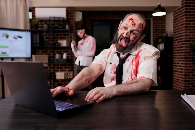 Zumbi assustador assustador usando laptop na mesa, parecendo assustador e horrível trabalhando no computador. Aterrador e dramático cérebro comendo monstros sentados no escritório de negócios, horrível caminhante sangrento.