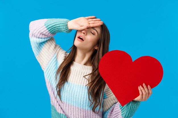 Zuneigung, Leidenschaft und Beziehungskonzept. Attraktive romantische Frau in der Winterstrickjacke, seufzend mit der Hand auf Stirn, nahe Augen und halten großes rotes Herz und verlieren ihren Verstand von der Liebe, blau