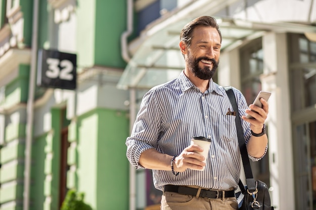 Foto zur arbeit gehen. positiver erwachsener mann, der lächelt, während er sein smartphone benutzt