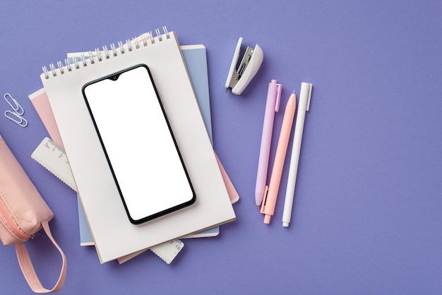 Zurück zum Schulkonzept Draufsichtfoto des Smartphones über einem Stapel Notizbüchern, Stiften, Heftklammern, Linealklammern und rosa Federmäppchen auf isoliertem violettem Hintergrund mit leerem Raum