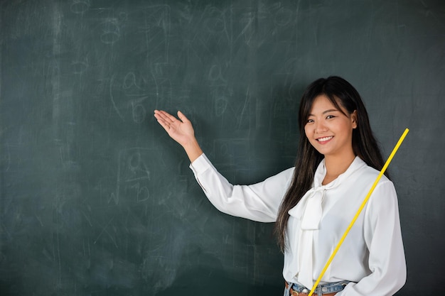 Zurück zum Schulkonzept. Fröhliche, schöne junge Frau, die mit dem Zeiger auf der Rückseite steht, asiatische Lehrerin, die mit Holzstab lächelt und auf die Tafel in der Schule im Klassenzimmer zeigt, Bildung