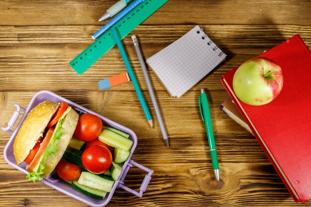 Zurück zum Schulkonzept Schulbedarf Bücher Apfel und Brotdose mit Burgern und frischem Gemüse auf einem Holztisch Draufsicht