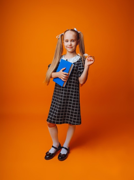 Zurück zur Schule Ein Schulmädchen hält ein blaues Buch vor einem isolierten gelben Studiohintergrund