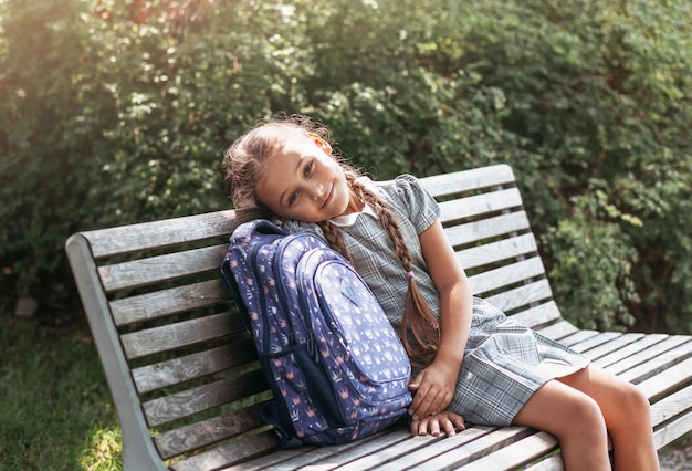 Zurück zur Schule Ein süßes kleines Schulmädchen in einem Kleid mit Zöpfen und großen blauen Rucksäcken sitzt auf einer Bank im Schulhof Ein kleines Mädchen geht in die erste Klasse