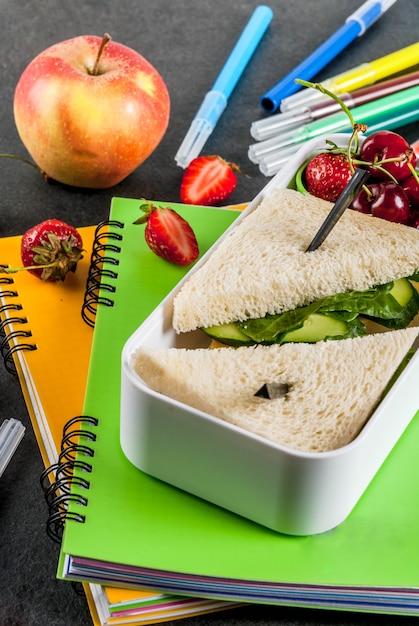 Zurück zur Schule. Eine herzhafte gesunde Schulmahlzeit in einem Kasten: Sandwiche mit Gemüse und Käse, Beeren und Früchten (Äpfel) mit Notizbüchern, Farbstifte auf einer schwarzen Tabelle.