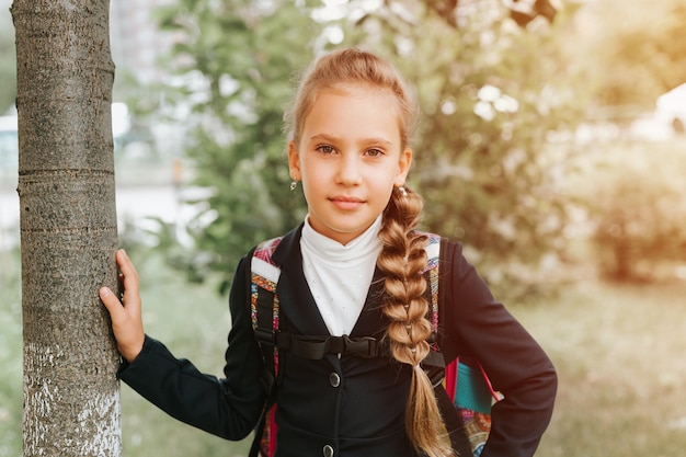 Zurück zur Schule kleines glückliches Kind Schülerin Schulmädchen acht Jahre alt in modischer Uniform mit Rucksack und Frisur voluminöser langer Zopf bereit, in die zweite Klasse am ersten Tag der Grundschule zu gehen