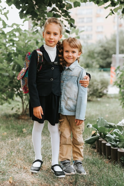 Zurück zur Schule kleines glückliches Kind Schülerin Schulmädchen acht Jahre alt in modischer Uniform mit Rucksack und ihrem Vorschulbruderjungen umarmen sich zusammen bereit für die zweite Klasse am ersten Tag der Grundschule