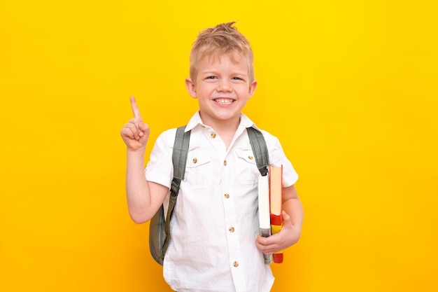 Zurück zur Schule Porträt der Grundschule des kleinen blonden Jungen mit Tasche und Büchern im weißen Hemd, das mit dem Finger nach oben zeigt Gelber Studiohintergrund Bildung Betrachtet und lächelt in die Kamera