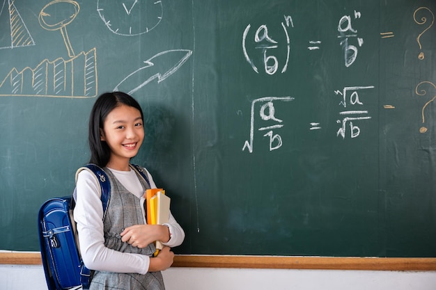 Zurück zur Schule. Porträt einer lächelnden Schülerin aus schwarzem Kreidebrett, glückliches schönes asiatisches Schulmädchen, das Bücher hält, die vor der Tafel des Klassenzimmers stehen, Unterrichtsstunde