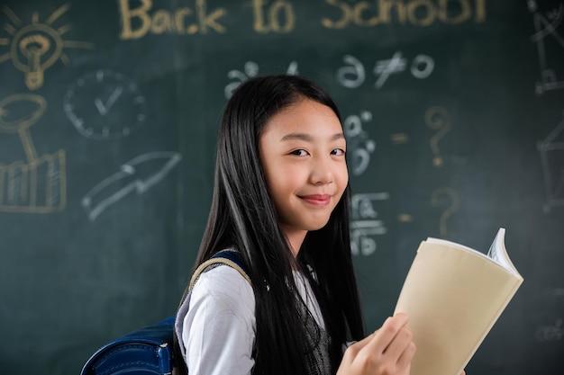 Zurück zur Schule. Porträt einer lächelnden Schülerin aus schwarzem Kreidebrett, glückliches schönes asiatisches Schulmädchen, das Bücher hält, die vor der Tafel des Klassenzimmers stehen, Unterrichtsstunde