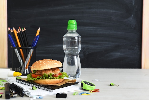 Zurück zur Schule. Quarantäne-Erziehung. Brotdose mit Hamburger, Wasser und Schulzubehör auf dem Hintergrund der Schulbehörde.