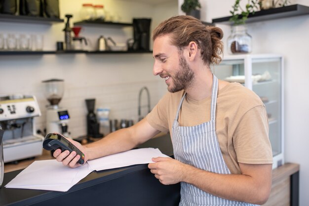 Zusammenfassen. Lächelnder junger erwachsener langhaariger Mann in der Schürze im Profil, das Rechnungen im Café prüft