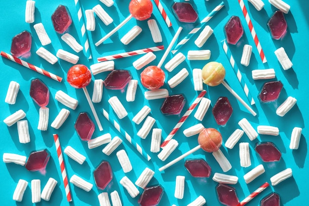 Zusammensetzung aus gestreiften Trinkhalmen, hellen Lutschern, luftigen Marshmallows und Stückchen kandierten Fruchtgelenks, die in einer bestimmten Reihenfolge vor blauem Hintergrund direkt über dem Blick liegen