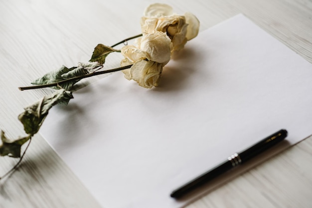 Zusammensetzung getrocknete Rosenblüten Blatt Papier und Stift für Nachricht auf weißem Hintergrund aus Holz Postkarte mit Kopierbereich Rahmen Platz für Textdesign Grußkarte Selektiver Fokus