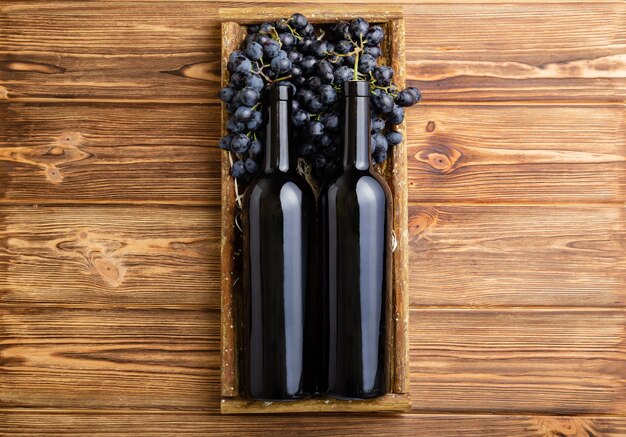 Zusammensetzung mit zwei Rotweinflaschen auf braunem Holztisch. Rotweinflaschen im Kasten auf schwarzen reifen Trauben auf Holztisch. Alte Sammlung Weinetikettenvorlage Ansicht von oben.