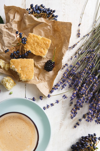 Zusammensetzung von Kaffee, Keksen, Beeren und Lavendelblumen, Nahaufnahme auf weißem Holz. Blaue Tasse mit cremigem Schaum