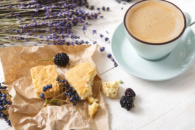 Zusammensetzung von Kaffee, Keksen, Beeren und Lavendelblumen, Nahaufnahme auf weißem Holz. Blaue Tasse mit cremigem Schaum