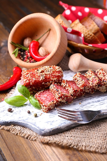 Zusammensetzung von Messer geschnittener Salamiwurst mit Sesam auf Schneidebrett auf Holzhintergrund