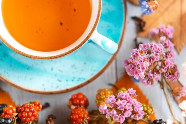 Zusammensetzungsstillleben eines Bechers mit heißem Blatttee mit Beeren und Herbstlaub auf einer Holzoberfläche