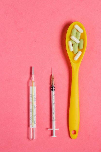 Zusammenstellung von Pillen und von Kapseln im gelben Löffel, Spritze, Thermometer auf rosa Hintergrund.