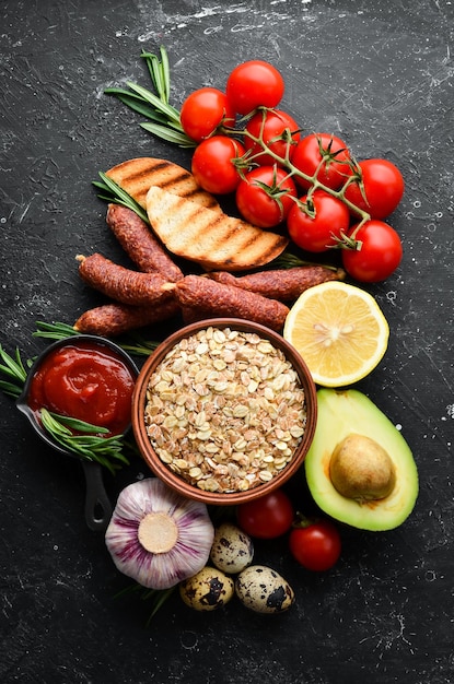 Zutaten einer gesunden Ernährung. Frühstückszubereitung. Eier, Haferflocken, Würstchen und frisches Gemüse auf schwarzem Steinhintergrund.