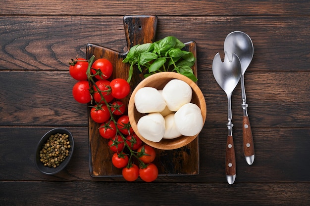 Zutaten für Caprese-Salat zum Kochen Schneiden von Holzbrettern mit traditionellen Caprese-Zubereitungszutaten Mozzarella-Tomaten-Basilikum-Olivenöl-Käse-Gewürzen auf rustikalem Holzhintergrund