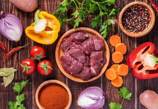 Zutaten für das Kochen von Gulasch oder Eintopf: rohes Fleisch, Kräuter, Gewürze, Gemüse auf dunklem Holzhintergrund.