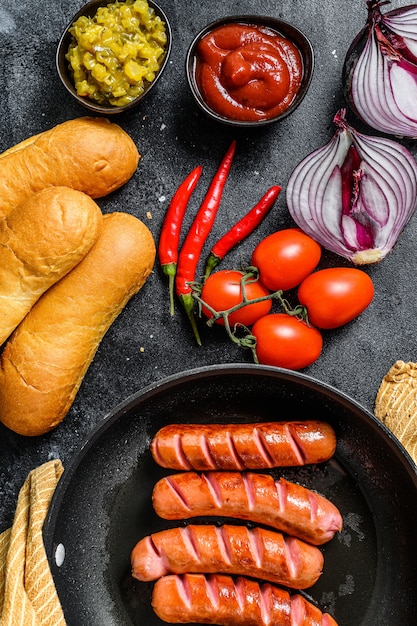 Zutaten für die Herstellung von hausgemachten Hot Dogs. Würstchen in der Pfanne, frisch gebackene Brötchen, Senf, Ketchup, Gurken. Schwarzer Hintergrund. Draufsicht